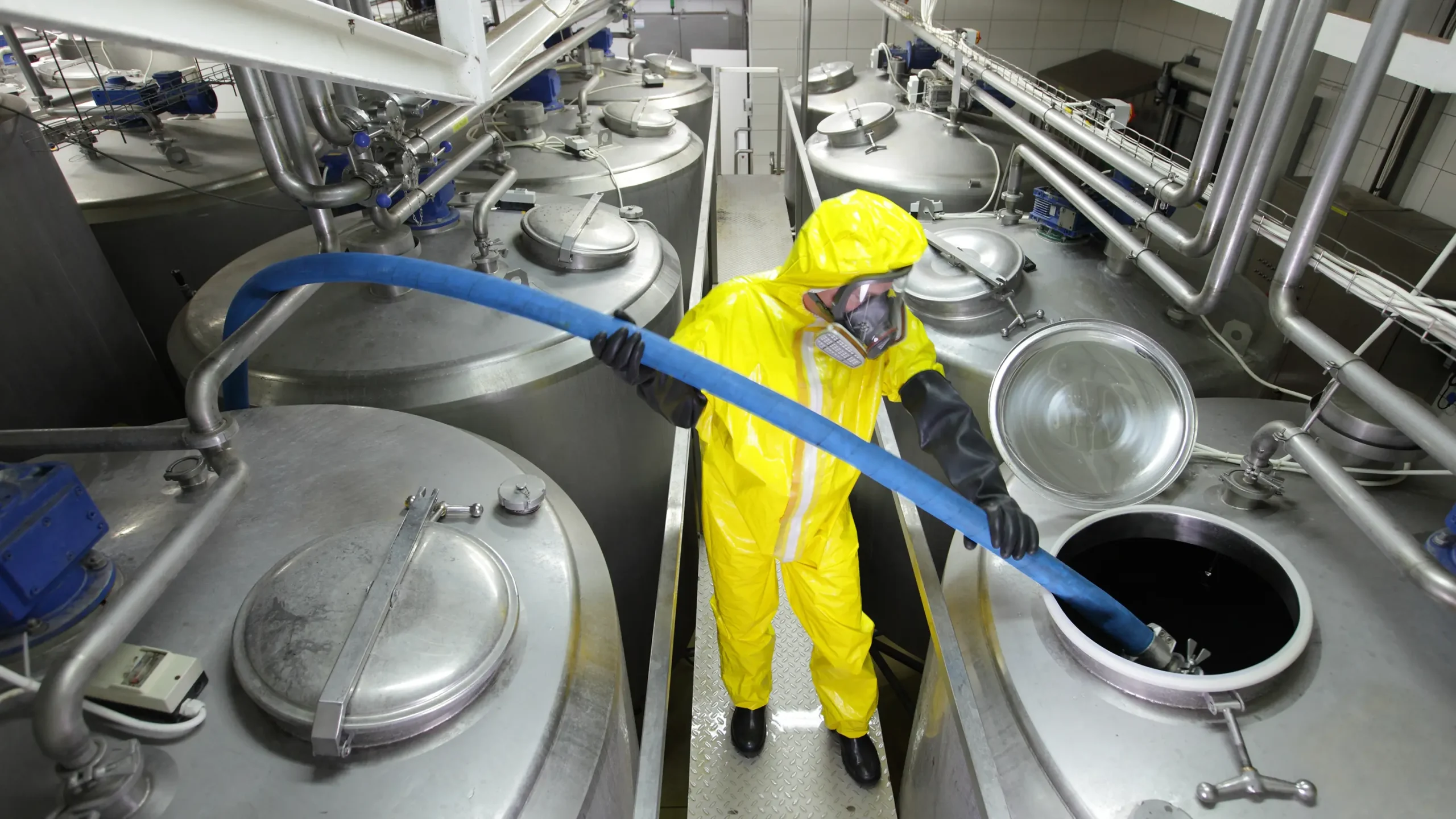 A worker in protective coveralls is seen pouring a substance into a large silver tank at a state-of-the-art facility for fine chemicals.