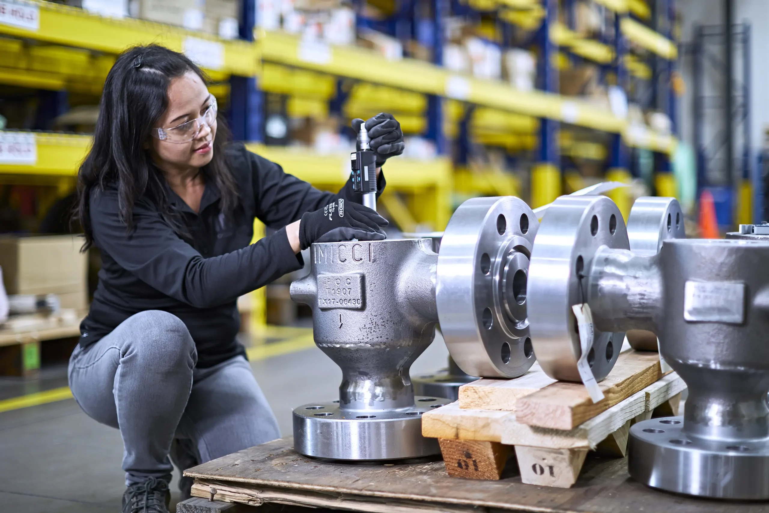 Woman in PPE inspects control valve at a workbench. Using tools, she examines for defects, records observations, and ensures quality control.